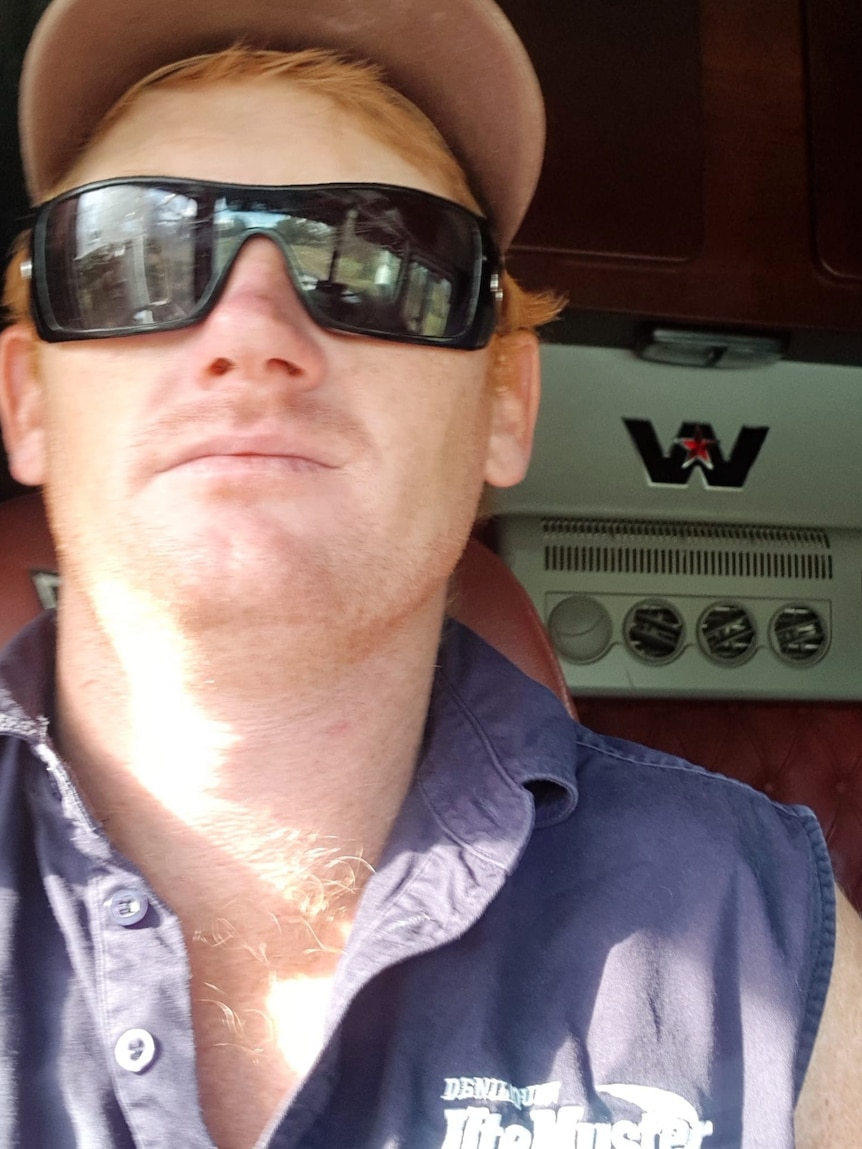 A man with ginger hair wearing sunglasses and a cap sitting in the cab of a truck.