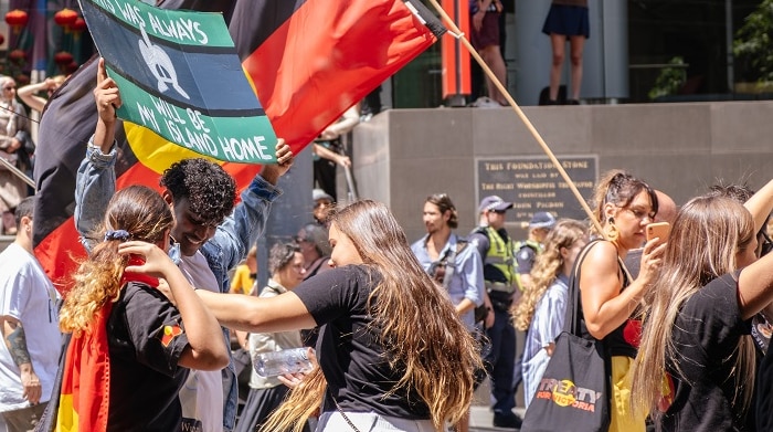A group of people are gathering on the street, waving Aboriginal and Torres Strait Island flags in a peaceful protest