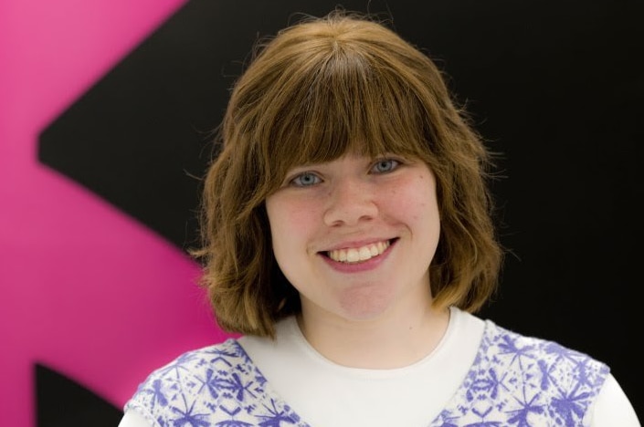 A picture of a woman smiling at the camera while wearing a purple dress. She stands in front of a pink and black background.