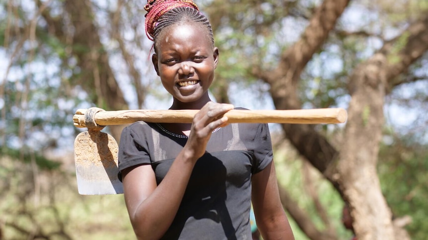 A young, smiling African woman holds a hoe over her shoulder