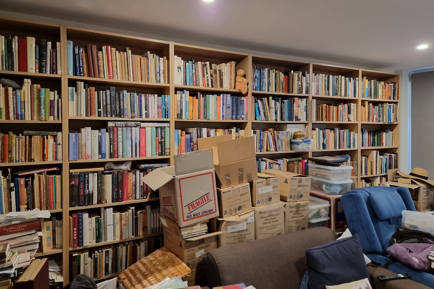 A living room with a packed bookshelf and various piles and boxes of books strewn around.