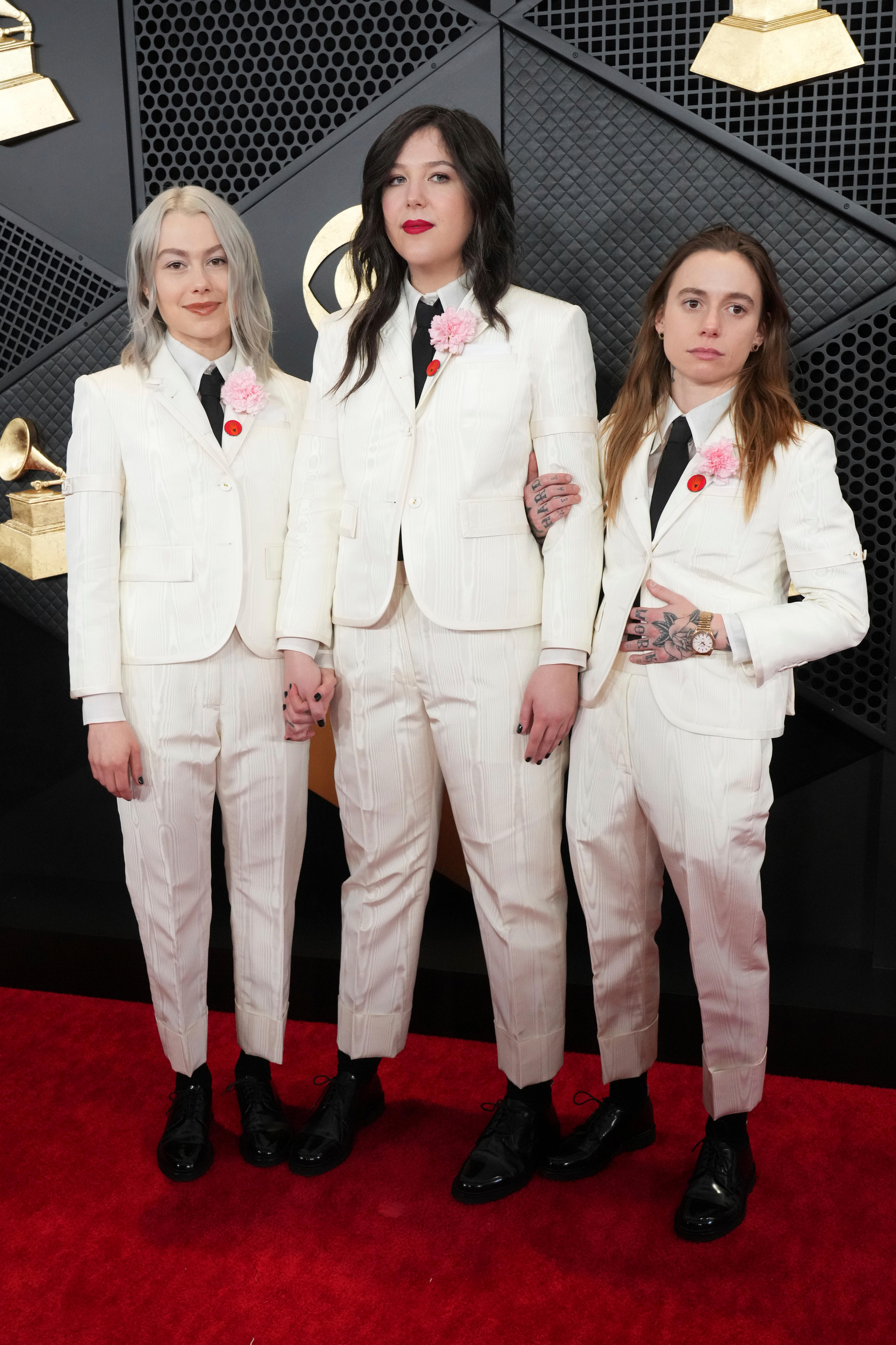 Phoebe Bridgers Lucy Dacus, and Julien Baker in white suits with black ties, each with a pink carnation and red dot on the lapel