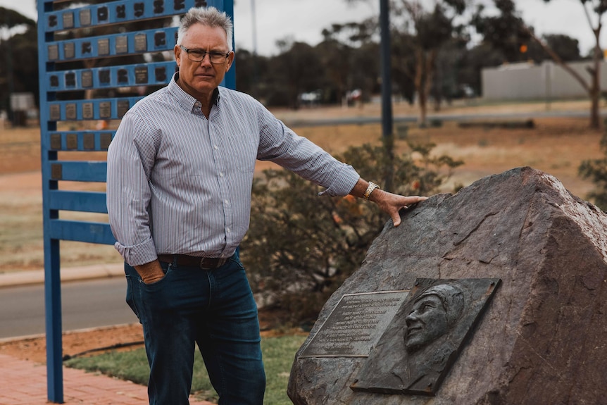 A man in a collared business shirt and jeans next to a memorial for a RFDS crew.     