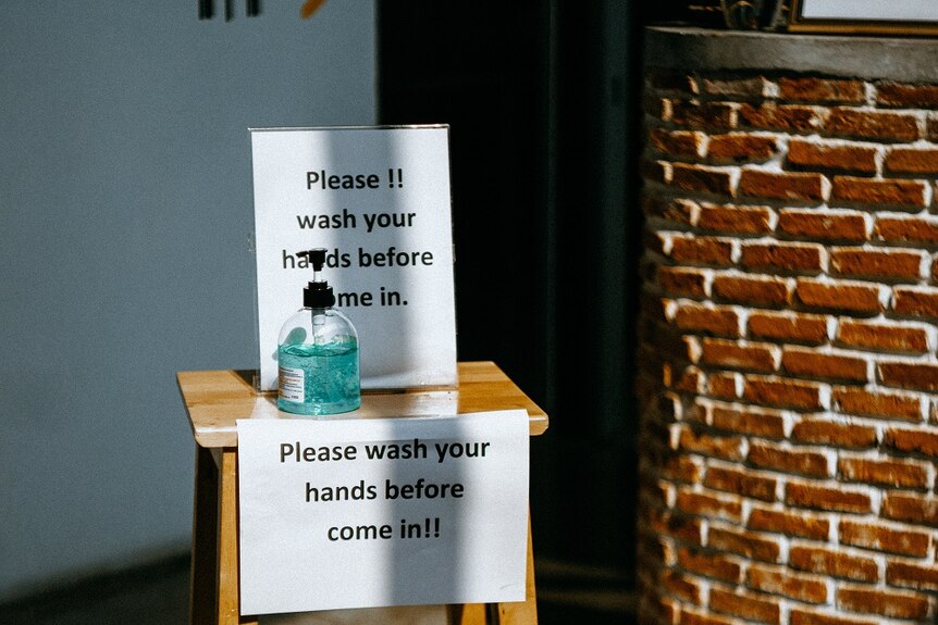 A bottle of hand sanitiser sits on a stool with a sign reading: "Please wash your hands before come in!!"