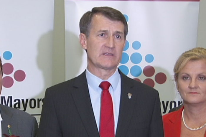 A man with dark hair, wearing a dark suit and a bright tie, addresses the media.