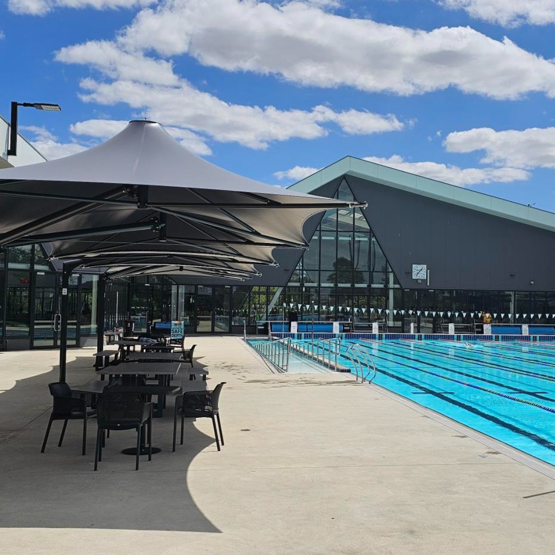 Tables and chairs on concrete are shaded by umbrellas beside a large pool.