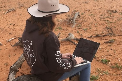 A rural student with her back to camera outside, on a laptop.