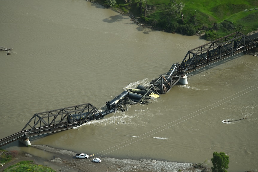 A birds eye shot shows a long metal bridge collapsed in a muddy river