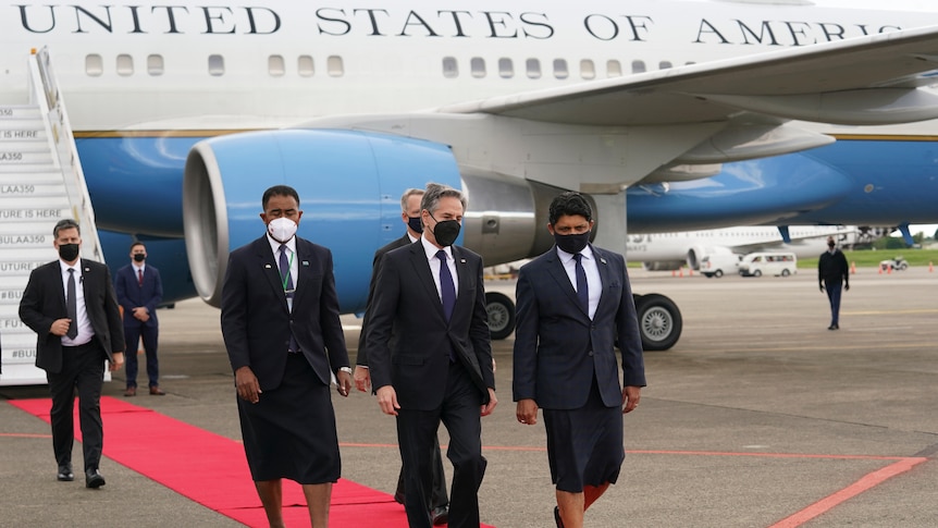 A man in a dark suit is flanked by two officials wearing Pacific formal dress as they walk down a red carpet away from a jet.