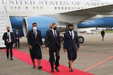 A man in a dark suit is flanked by two officials wearing Pacific formal dress as they walk down a red carpet away from a jet.