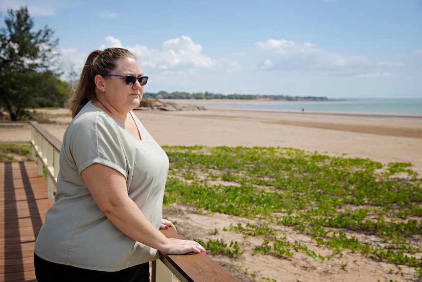 A woman wearing sunglasses leans on a handrail and looks out onto a beach. 