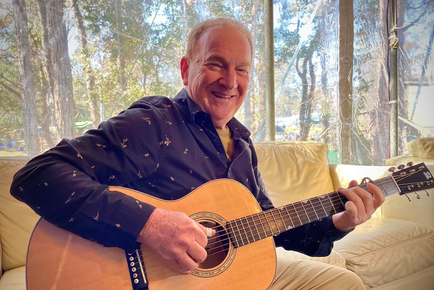 A man sits on a couch holding a guitar smiling.