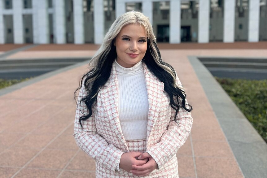 A young woman with dyed black-and-white hair stands in front of Parliament House, dressed in a pink suit jacket.