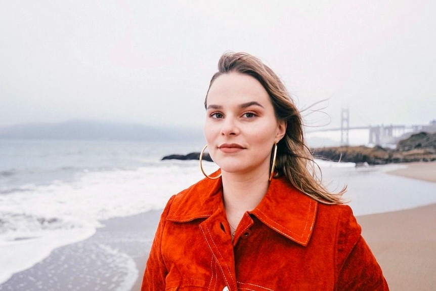 A woman with long brown hair wearing a red coat stands on a beach.