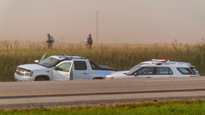Two policeman stand in a field near a large SUV with its doors open, and two large police vehicles, near a road.
