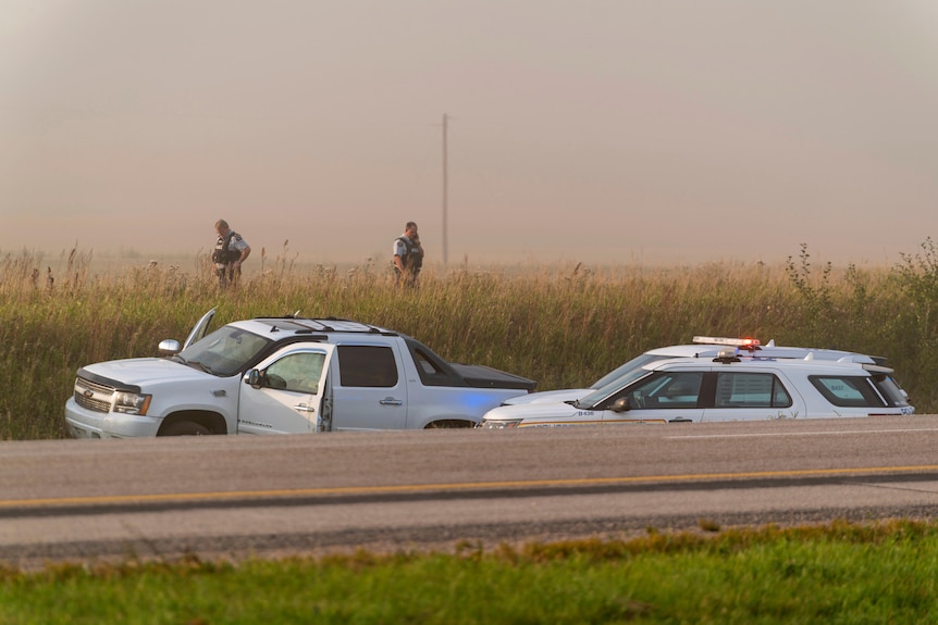 Two policeman stand in a field near a large SUV with its doors open, and two large police vehicles, near a road.