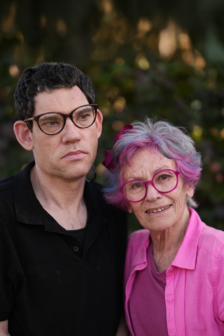 A middle-aged man and an elderly woman with pink hair and glasses stand close together outdoors.