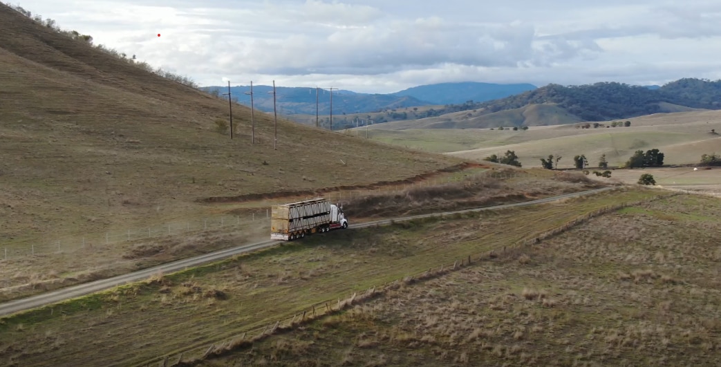 A double decker cattle truck drives down a dirt road, with green paddocks on either side, looking down a hilly valley.