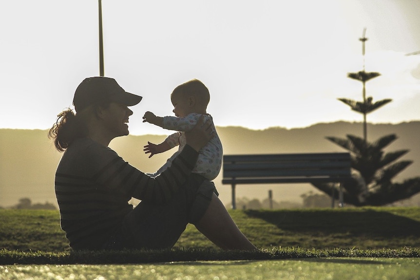 A woman sits down in a park and holds up her baby and they are smiling at each other.