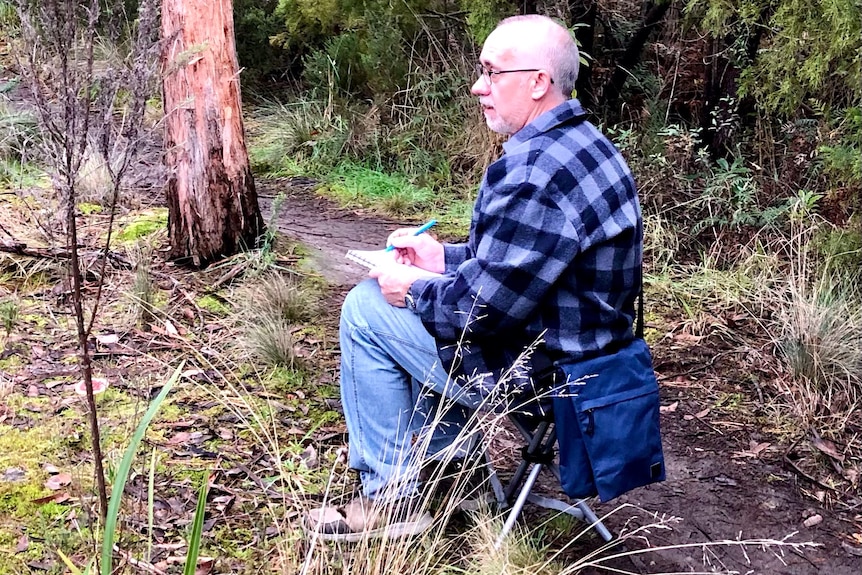 a man in a blue checked shirt is sitting in the bush whole holding a journal and pen