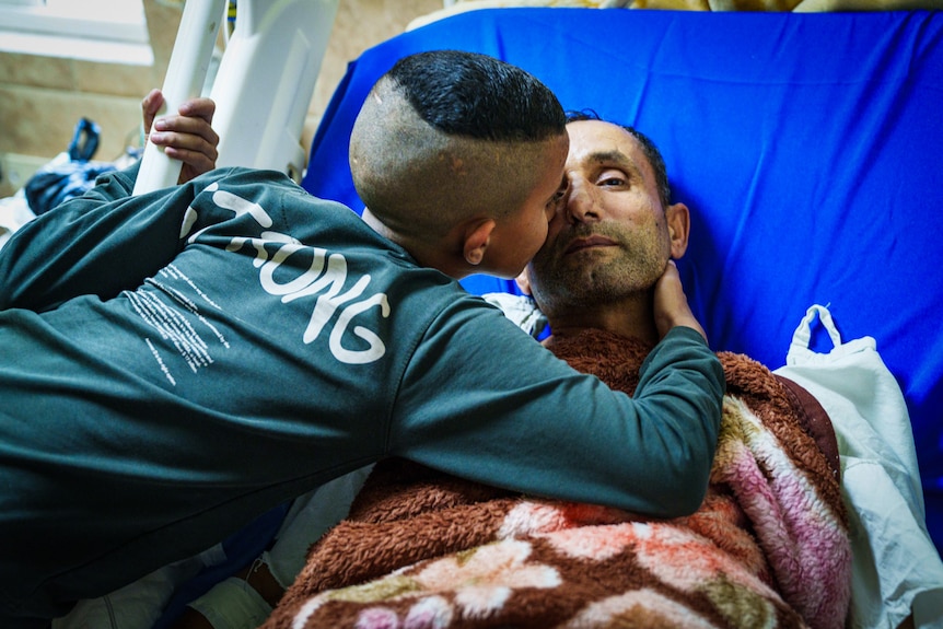 A man lies in a hospital bed wrapped in a blanket as he is kissed by his son.