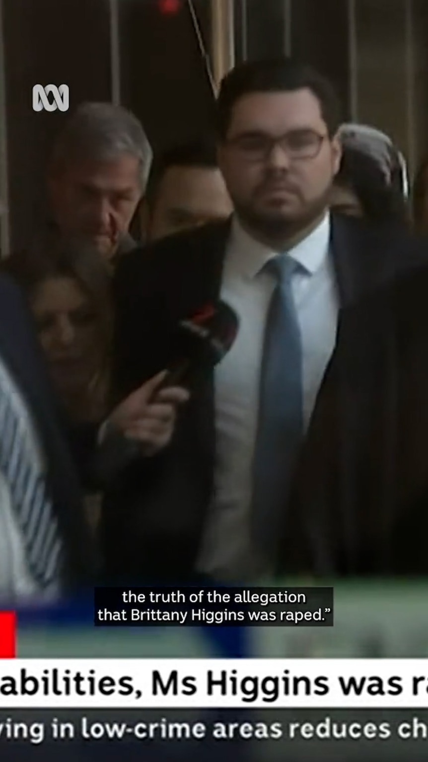 Man walks unsmiling while surrounded by journalists with microphones and cameras