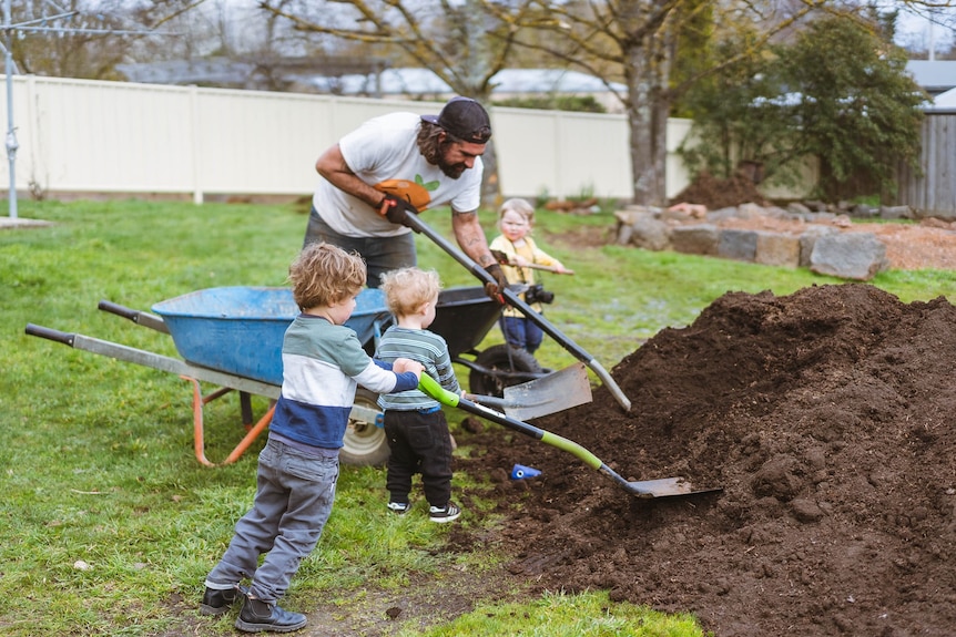 A man and children with shovels, working on a mound of soil, with a nearby wheelbarrow.