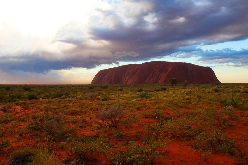 distant view of Uluru against red soil, green vegetation, storm clouds above