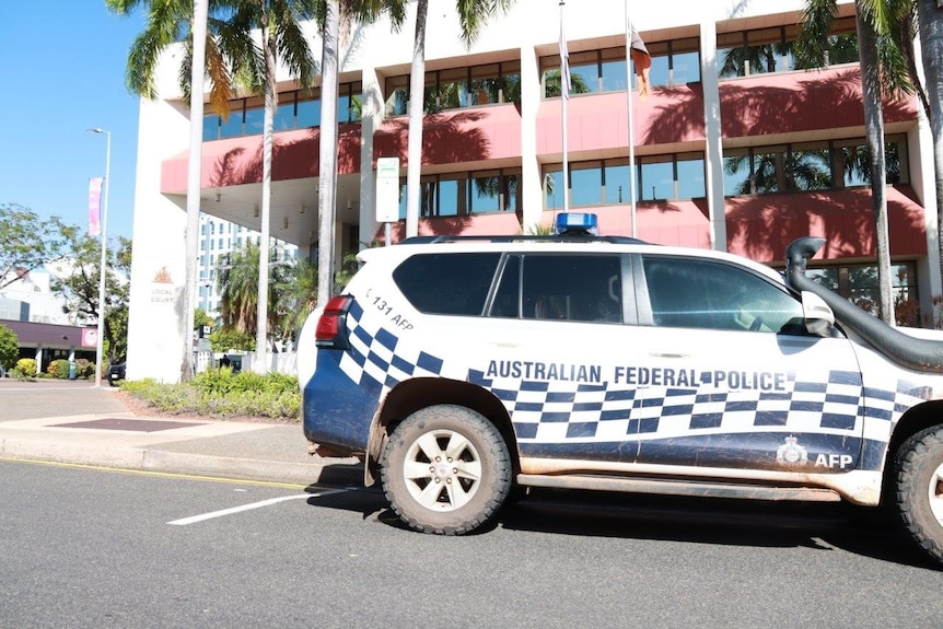An Australian Federal Police Officer with splotches of red dirt is parked under palm trees outside the court house