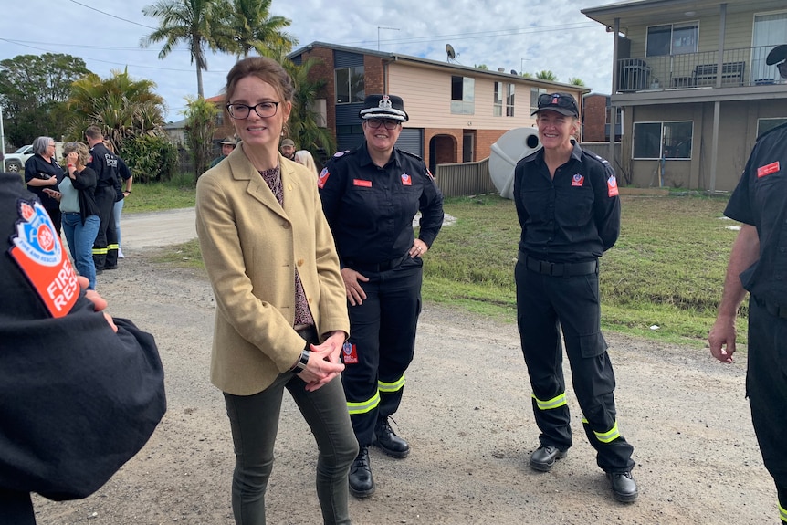 A woman with glasses and beige jacket on a residential street with firefighters.
