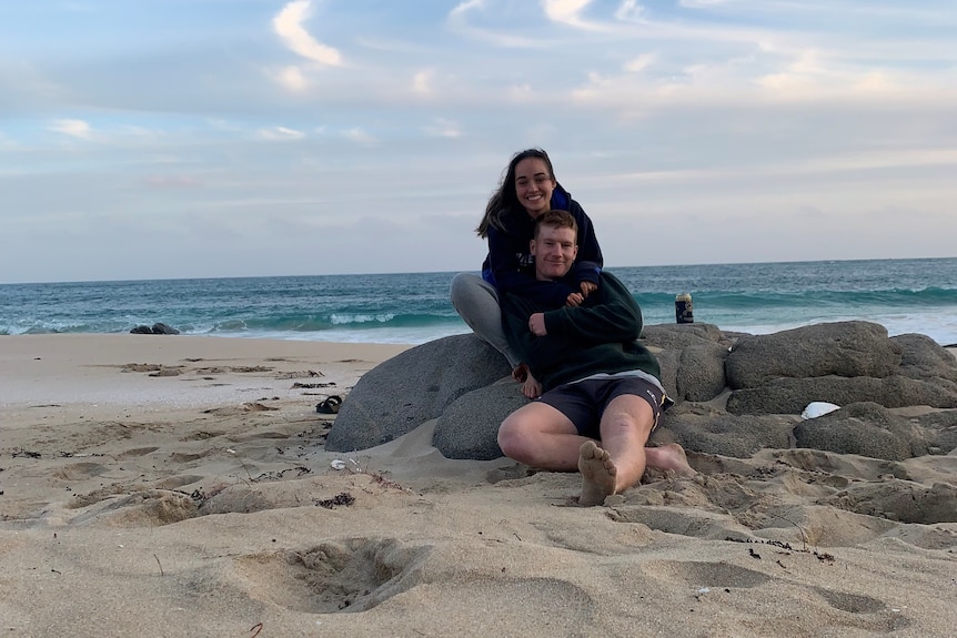 Hannah and her partner sitting close together on a beach, with sand and the ocean visible, in the evening.
