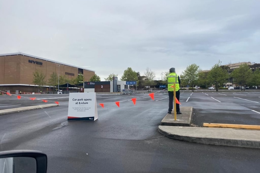 A security guard next to orange bunting at a car park with sign saying "Car park opens at 8:45am"