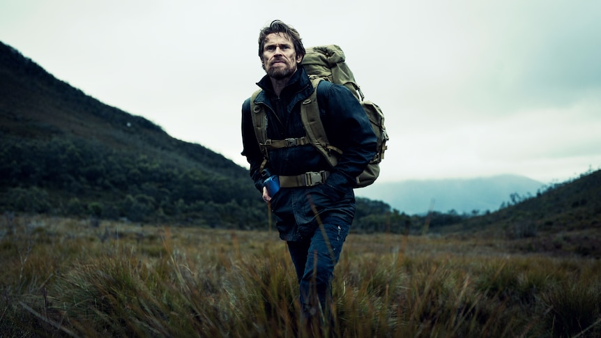 Willem Dafoe in The Hunter. He has a large backpack on and is standing alone in a grey Tasmanian landscape.