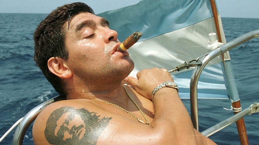 Shirtless Diego Maradona leans back on a boat smoking a cigar with an Argentinian flag behind.