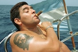 Shirtless Diego Maradona leans back on a boat smoking a cigar with an Argentinian flag behind.