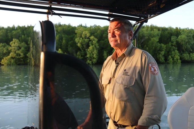 A man looks off into the distance, he is on a boat and there are mangroves in the background.