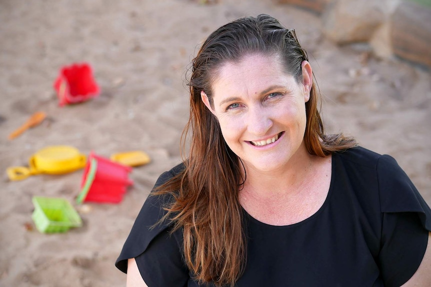 A woman with brown hair and a black top smiles, with a sandpit and children's toys behind her.