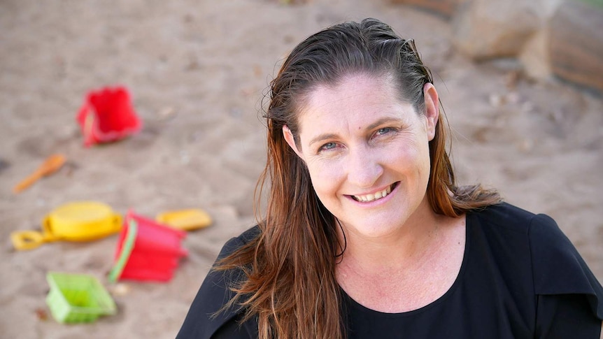A woman with brown hair and a black top smiles, with a sandpit and children's toys behind her.