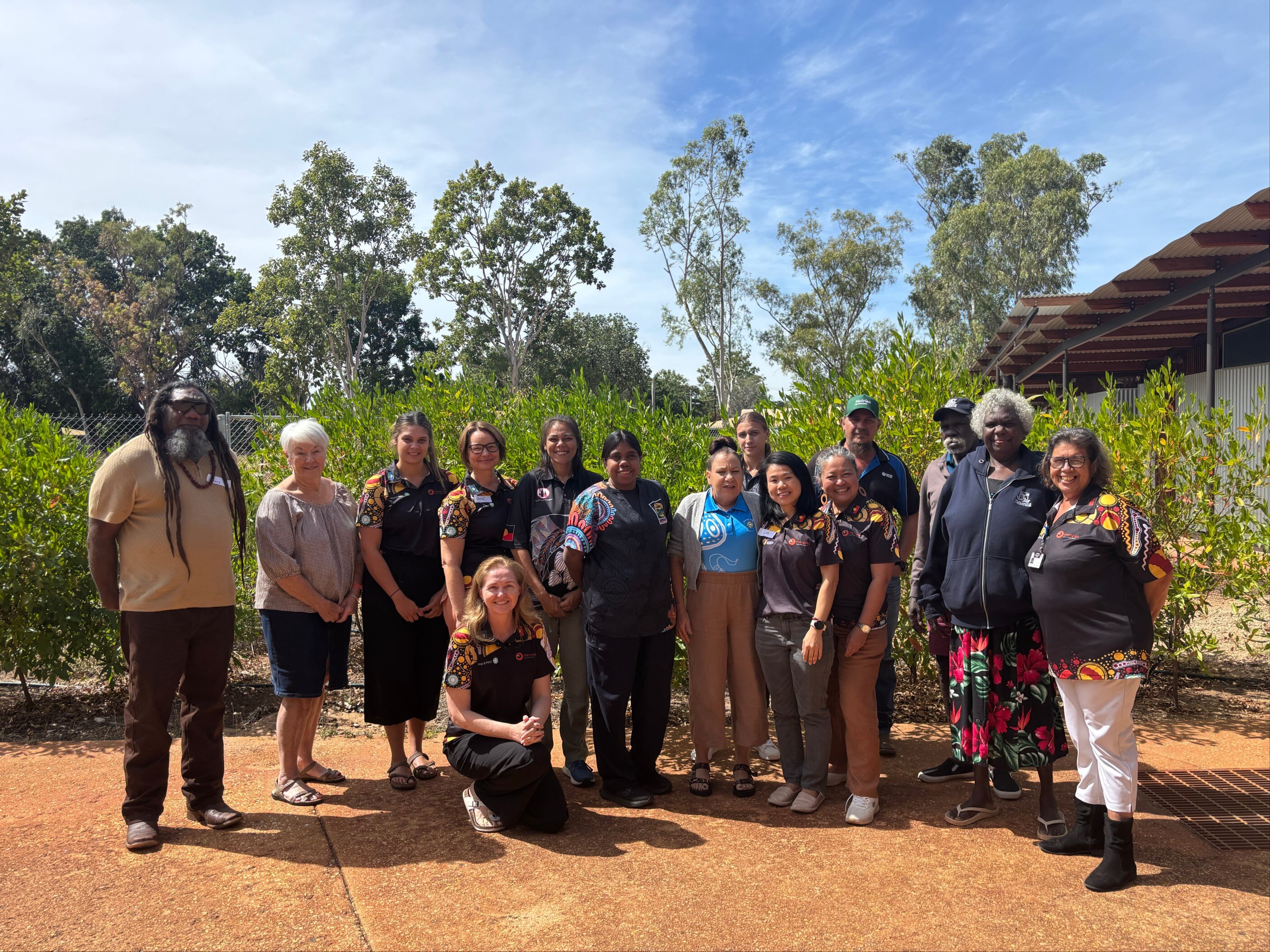 15人のグループ、先住民族と非先住民族のグループが、ブッシュの前の赤い汚れにグループ写真のために集まりました。