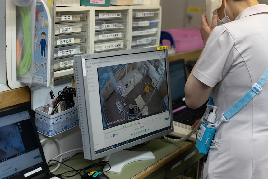 A woman in a white nurse's uniform on the phone stands in front of a baby video monitor 