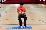 Que Jianyu sits in a chair in a gymnasium with a Rubik's cube in each hand and one at his feet. A woman is timing him.