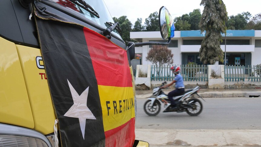 Red, yellow and black flag with a white star on the front of a bus with a motorcyclist going past in the background.