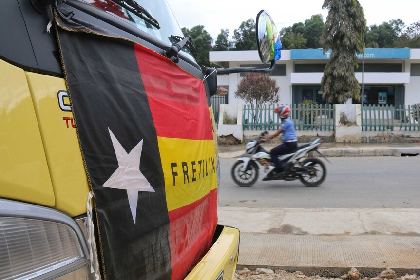 Red, yellow and black flag with a white star on the front of a bus with a motorcyclist going past in the background.