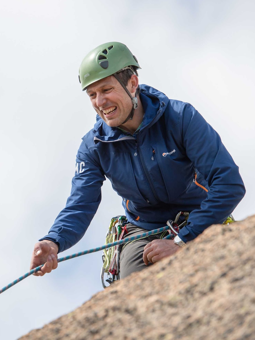 Man wearing green helmet stands on a rock on a mountain holding a rock climbing rope.