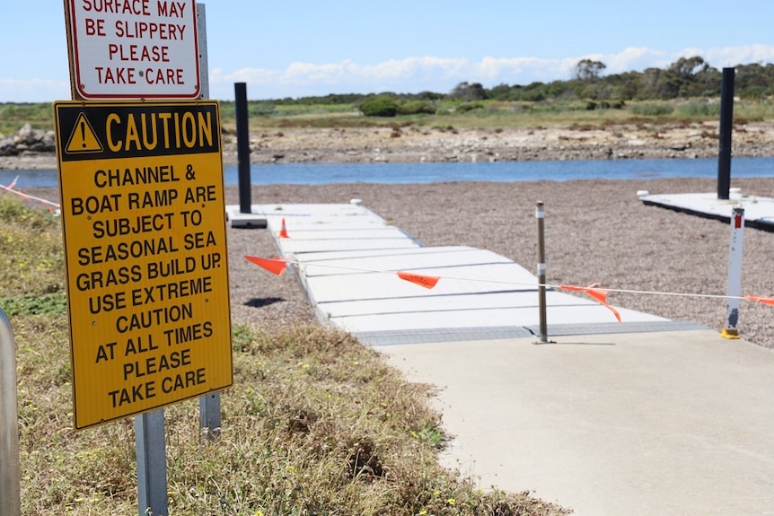 A "caution" sign in front of a boat ramp leading to sand. Orange flags strung across the entrance.