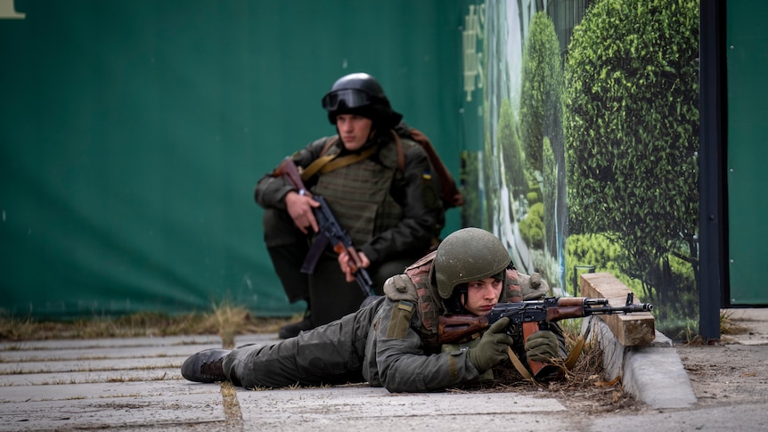 Two Ukrainian soldiers, one crouching and one lying while looking through the scope of a gun.