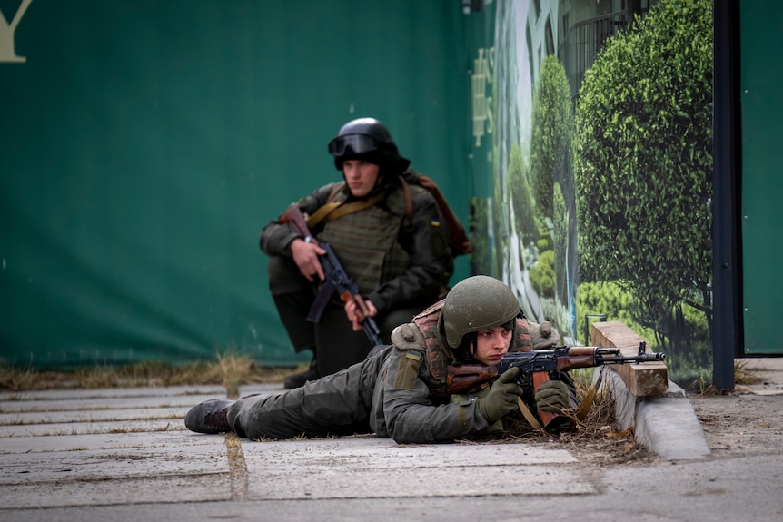 Two Ukrainian soldiers, one crouching and one lying while looking through the scope of a gun.