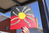 A child's hand holds up a painting of the Aboriginal flag with leaves stuck to it.