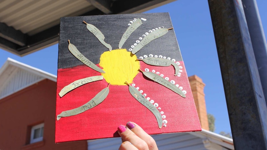 A child's hand holds up a painting of the Aboriginal flag with leaves stuck to it.