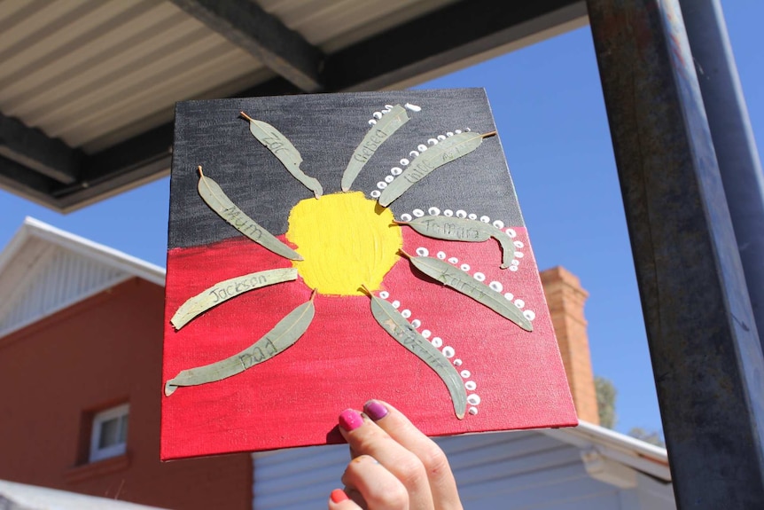 A child's hand holds up a painting of the Aboriginal flag with leaves stuck to it.